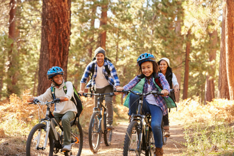 A smiling young family riding their bikes down a wooded path.