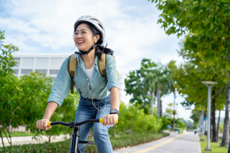 Smiling Asian woman in her 20s wearing casual clothes and headphones riding a bicycle on the sidewalk in the park. Look to the side. The concept of healthy living in the city.