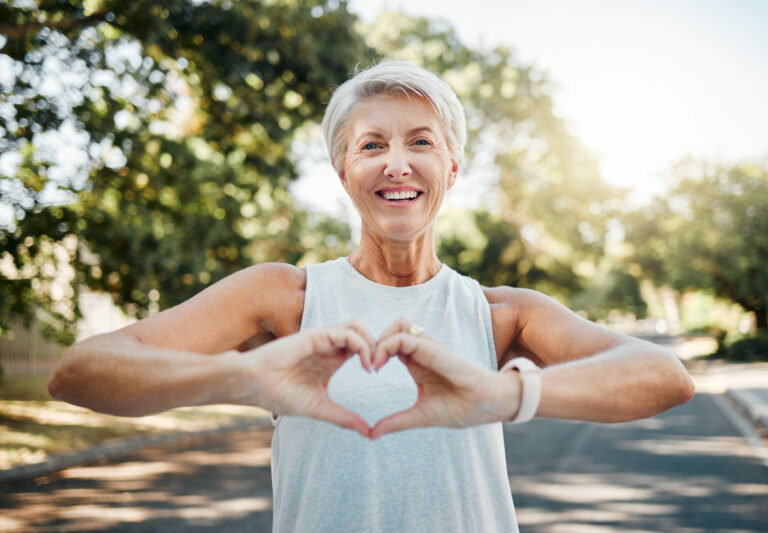 Fitness, happy and heart hands of senior woman in nature after running for health, wellness and workout. Smile, motivation and peace with senior lady and sign for love, faith and training in nature